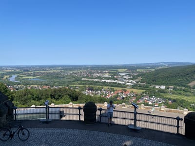 The image shows a panoramic view from a vantage point in Porta Westfalica, Germany. In the foreground, there is a paved area with cobblestones and a black bicycle parked on the left. A black metal railing encloses a walkway or observation deck. A man wearing a white t-shirt, blue shorts, and a helmet stands near the railing, looking towards the right. He is holding something in his hands. Two other people are visible in the background, further down the walkway.

Beyond the railing, the scene opens up to a sprawling landscape. A town with numerous houses, predominantly with red-tiled roofs, is nestled among lush green trees. A river or body of water winds through the landscape, with bridges crossing it in the distance. Further out, the land stretches towards a hazy horizon, with rolling hills and a clear blue sky above. Various structures, including industrial buildings and wind turbines, are scattered throughout the town and surrounding areas. The overall impression is a sunny day with excellent visibility, offering a commanding view of the city and its environs.
