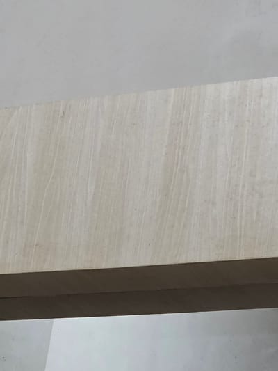 The image displays a close-up view of a light-colored, textured surface, possibly wood or stone, with vertical striations. The surface is illuminated from above, creating subtle shadows that enhance the texture. A darker, horizontal band is visible along the bottom edge of the frame, suggesting the underside of an object or architectural feature. The background above the textured surface is a plain, light gray. This image was taken in Bireuenmeunasahblang, Indonesia, in an indoor or sheltered environment.