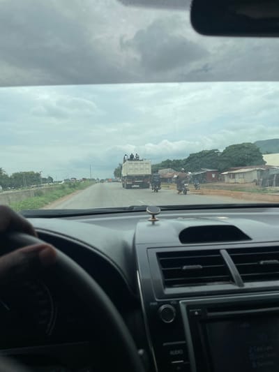 This media file captures a view from the interior of a vehicle, likely a car, as it travels along a road. The scene outside the windshield depicts a rural or semi-urban setting in Kwali, Nigeria.
The primary subjects visible are a series of vehicles on a road, including a large white truck in the foreground loaded with passengers, several motorcycles, and other cars further down the road. There are also buildings and trees lining the road. The sky is cloudy, suggesting overcast weather, and the overall lighting indicates it is daytime. The perspective suggests the vehicle is moving, contributing to a sense of travel.
Notable details include people riding on top of the white truck, which is a significant visual element. The dashboard of the vehicle is partially visible, showing an air conditioning vent and part of the audio system. The road itself appears to be paved, with a grassy verge.
There is no visible text in the image. The activity taking place is vehicular movement along a road, likely a common travel route in the area. The image conveys a sense of everyday life and transportation in the depicted location.