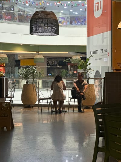 The image depicts an indoor setting, likely a food court or common area within a commercial establishment. In the foreground, to the right, are several green plastic chairs. The floor is polished and reflects light.
In the mid-ground, a man and a woman are seated at a table. The woman, with dark, curly hair, is wearing a light-colored dress and is facing away from the camera. The man, wearing a grey polo shirt and dark pants, is seated to her right, with his legs crossed. Both appear to be dining or resting. Between them, a large, textured pot contains two green plants.
Behind the diners, a glass partition separates them from the rest of the establishment. Through the glass, a restaurant with a sign reading "bendito BURRITO" is visible. Further in the background, on the left, is a rack of flags. On the right, a large banner is prominently displayed, featuring the "Xiaomi 17T Pro" logo and text. This banner suggests a retail or promotional area within the location. The ceiling features decorative lighting fixtures, including a large, woven pendant lamp in the upper left quadrant. The overall environment suggests a modern, commercial space. The specific location within Perímetro Urbano Barranquilla, Colombia cannot be determined from the visual information alone.