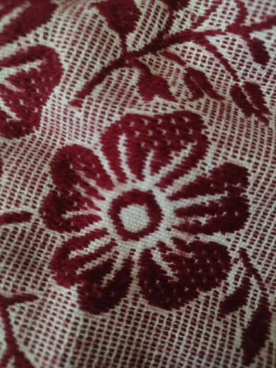 This media file is a close-up, overhead shot of a woven fabric with a floral pattern in red and white. The texture of the fabric is a prominent feature, showing a grid-like weave with raised threads forming the design.
The main subject is a stylized red flower with distinct petals. The flower is centrally located in the frame, with a smaller, white center. Around the flower, red leaf-like shapes and swirling vine elements are visible, all rendered in the same textured, woven style against the white background weave. The pattern appears to be repetitive, suggesting it might be part of a larger textile like a tablecloth or curtain.
There are no people, specific activities, or events depicted. The lighting appears to be natural and diffused, casting no harsh shadows and suggesting an indoor setting or an overcast day. There are no visible location cues, time of day indicators, or emotional elements. No text is present in the image. The image captures a detail of a decorative textile, likely from a home setting, given the context of Țipordei, Moldova, where such fabrics are often used for home furnishings.