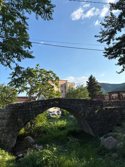 The image depicts an old stone arched bridge in Podostrog, Montenegro. The bridge is made of rough-hewn stones and is covered in some greenery. Beneath the arch, a small stream or dry creek bed is visible, with lush green grass and rocks. On the other side of the bridge, several cars are parked on a paved area. In the background, there is a multi-story building with a light-colored facade, and further behind that, rolling green hills under a clear blue sky with scattered white clouds. Trees with green foliage frame the top of the image, and power lines are strung across the sky. A sign for "MAINE Night Club" is visible on the right side of the bridge's railing. The scene appears to be a quiet, outdoor setting, likely during the daytime.