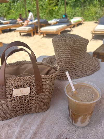 On a sandy beach in Chernomorets, Bulgaria, a woven straw bag sits to the left of a wide-brimmed straw hat. In front of the hat and to the right of the bag is a clear plastic cup filled with a frothy, layered beverage, likely coffee, with a white straw. In the background, several empty lounge chairs are visible under umbrellas, with people seated on some of them. Lush green foliage is present behind the beach setup. The scene suggests a relaxed day at the beach.