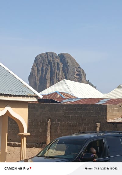 The image depicts a mountainous landscape in Wase, Nigeria, set against a clear blue sky. In the foreground, urban development is evident with residential buildings exhibiting a mix of corrugated metal roofing. A tan-colored structure with an arched entrance is partially visible on the left. A dark-colored SUV is positioned at the bottom of the frame, with a person visible in the driver's seat. The photograph is taken with a Camon 40 Pro, with a focal length of 78mm at f/1.8, a shutter speed of 1/2278 seconds, and an ISO of 52.