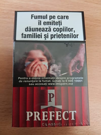 The image shows a close-up of a pack of "PREFECT CLASSIC" cigarettes. The pack has a striking design featuring a red and white striped background on the right side, with bold white lettering spelling out "PREFECT". On the left side, a portion of a health warning is visible, written in Romanian. The text translates to "The smoke you emit harms children, family, and friends." Below this, there's a further message in smaller print: "To obtain information about smoking cessation programs, call 0800-10001 or visit www.imspdrn.md".
The center of the pack displays a stark image of a person, likely a child, with their hands covering their mouth and nose, suggesting distress or suffocation from smoke. A hand holding a lit cigarette is also visible in the upper part of this central image, with wisps of smoke curling around it.
The lighting in the image is neutral, and the setting appears to be indoors, possibly on a wooden surface. The overall impression is one of a public health warning campaign, highlighting the detrimental effects of smoking, particularly on vulnerable individuals. The "Kamenka, Moldova" location context suggests this pack is intended for the Moldovan market.