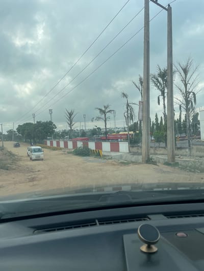 The image shows an outdoor scene in Karmajiji, Nigeria, on a cloudy day. The perspective is from inside a vehicle, looking out at a dirt road lined with power poles and wires. Palm trees with sparse fronds are visible along with other greenery. In the background, there are buildings with red and white striped facades, suggesting an industrial or potentially an airport-related area. A white truck and another smaller dark vehicle are on the dirt road. The overall mood is overcast and perhaps a bit hazy. There is no visible text in the image.