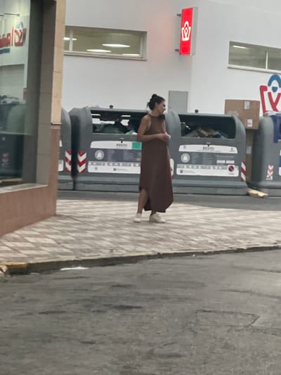 An adult female, with dark hair pulled back into a bun, stands on a tiled sidewalk in front of a row of large, grey recycling bins. She is wearing a long, brown sleeveless dress and white sneakers. The bins are partially full of various items, including what appears to be cardboard boxes and plastic. Above the bins, on a white wall, is a red sign with a white shopping bag logo. To the left of the bins, part of a building facade with a window is visible. The ground in front of the sidewalk is asphalt. The image appears to have been taken in La Línea de la Concepción, Spain, given the context provided by the prompt.