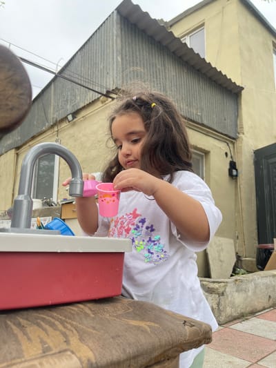 The image shows a young girl playing with a toy sink outdoors. She is wearing a white t-shirt adorned with colorful sequins. The toy sink is pink and grey, and it appears to be a miniature replica of a kitchen sink. The girl is holding a small pink cup up to the faucet, as if she is filling it with water. Her expression is one of concentration and engagement with her play.

The background suggests an outdoor setting, possibly a backyard or courtyard. There is a rustic, weathered building with a corrugated metal roof visible behind the girl. The building's walls are a pale yellow color. The ground in the foreground is paved with tiles in a pattern of pink and beige squares. The lighting suggests it might be daytime, possibly overcast.

The overall scene depicts a child engrossed in imaginative play, using a toy to mimic everyday activities. The environment appears modest and lived-in, hinting at a domestic setting in Sabunchu Raion, Azerbaijan. There is no visible text in the image.