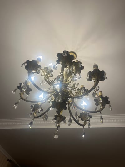 This media file shows a close-up, upward-looking shot of a decorative chandelier. The chandelier is made of what appears to be aged brass, with ornate, leaf-like accents and dangling crystals that catch the light. Several light bulbs are visible, illuminating the fixture and casting light patterns on the white ceiling. The chandelier has multiple arms extending from a central point, creating a multi-tiered effect.
In the background, a white decorative molding or cornice runs along the edge of the ceiling, suggesting an interior setting, likely a room with classical or traditional decor. The lighting suggests it is either nighttime or the room is dimly lit, with the chandelier being the primary light source. The overall mood conveyed is one of elegance and traditional opulence. There is no text visible in the image. The location context is Edem, Russia.
