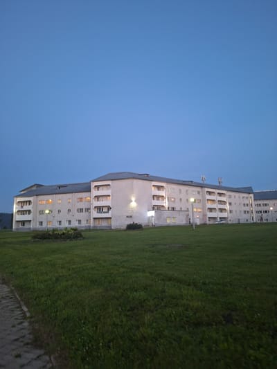 The image depicts a long, multi-story apartment building in the city of Atamanovka, Russia, under a twilight sky. The building is light-colored with a gray roof and features numerous windows, some of which are illuminated, suggesting occupancy. The architecture is functional, with balconies and some circular window elements. In front of the building, there is a well-maintained green lawn with a small landscaped garden bed in the foreground. A paved walkway runs along the left edge of the frame. The scene appears peaceful, with no overt signs of activity beyond a few indistinct figures visible near the building. The overall impression is that of a residential area at dusk.