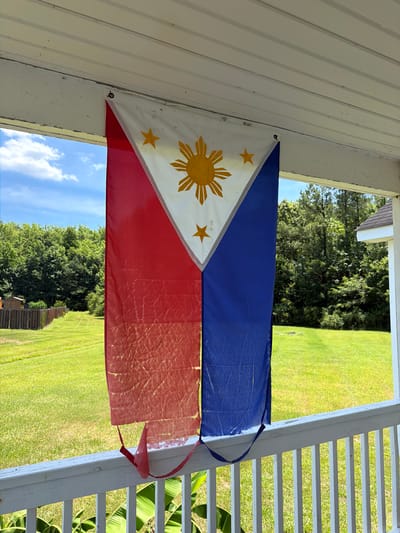 A Philippine flag hangs on a white porch ceiling. The flag has a red and blue vertical band, with a white triangle on the hoist side containing a yellow sun and three stars. The flag appears to be made of a thin material and has some creases and wear. Behind the flag, a green grassy yard extends towards a line of trees under a bright blue sky with scattered clouds. A wooden fence borders the yard. The porch railing is white and weathered. The scene is outdoors, suggesting a residential area in Elizabeth City, United States.