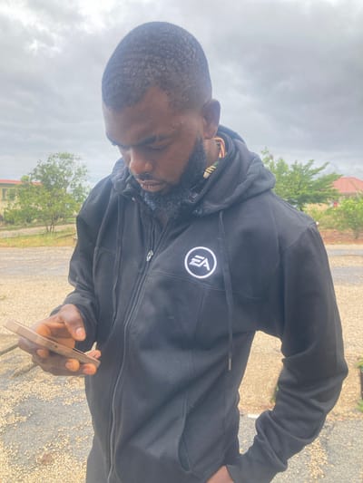 The image captures an outdoor, slightly low-angle shot of a man under an overcast sky. The man, who appears to be of African descent, has a short, dark haircut and a full beard. He is wearing a black zipped hoodie with a white circular logo on the left chest, which clearly displays the letters "EA". Near his right ear, what appears to be an earbud or earphone is visible.
He is holding a light-colored smartphone in his right hand and is looking down intently at its screen, suggesting he is actively using it. His expression is focused and neutral.
The setting appears to be a somewhat rural or undeveloped outdoor area in Bisichi, Nigeria, given the context. The ground beneath him is a mix of gravel and dirt. In the background, green trees are visible, and a building with a reddish roof can be partially seen through the foliage. The sky is largely covered with white and grey clouds, indicating an overcast day with soft, diffused natural light. The image itself is oriented vertically, rotated 90 degrees clockwise, making the man appear to be lying on his side or looking upwards from a prone position in the original orientation.