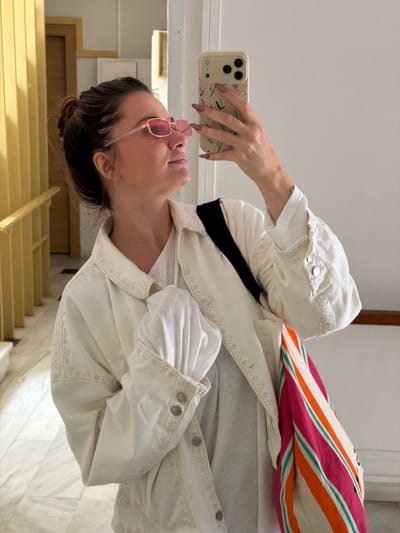 A woman with brown hair pulled back in a bun is taking a selfie with her smartphone. She is wearing pink-tinted, square-framed glasses and a white jacket over a white shirt. The jacket has a black strap or detail visible near her neckline. In her left hand, she is holding a tan-colored smartphone with a floral-patterned case and long, pink-colored nails. The background appears to be a building interior with a staircase. The steps are light yellow, and there is a doorway on the left side of the frame. The environment seems clean and well-lit, with marble or a similar type of stone visible on the floor and walls. Given the location in Glifada, Greece, it is likely that the photo was taken in an interior space such as an apartment building, office, or hotel.