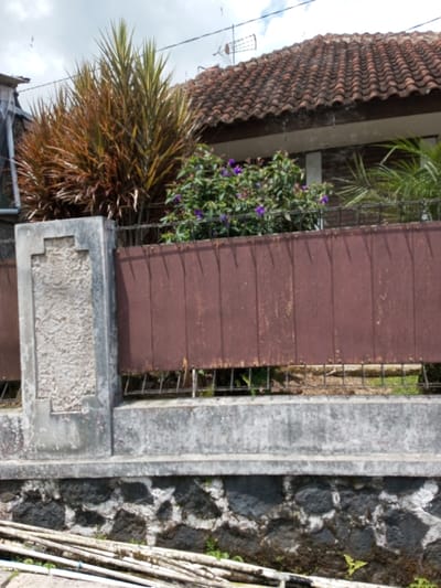The image depicts an exterior scene within the city of Viengthong, Laos, focusing on a property boundary. In the immediate foreground, several light-colored cylindrical objects, possibly pipes or bamboo, lie horizontally on disturbed ground. Below these objects is a wall constructed of dark, irregularly shaped stones mortared with a lighter material, from which small green plants emerge.

Dominating the midground is a fence structure. Its base consists of a light grey concrete retaining wall. Above this, the fence transitions into a section composed of vertical, dark reddish-brown panels, likely wood or metal, which are affixed between light grey concrete posts. The leftmost visible concrete post features a decorative, recessed rectangular panel with a cross-like design in textured lighter grey material. A dark metal grid is visible beneath the reddish-brown panels, above the concrete base.

Behind the fence, varied vegetation is prominent, including two large plants with reddish-brown and green variegated leaves, and a bush displaying numerous small, vivid purple flowers. Further in the background, a building is partially visible, identifiable by its reddish-brown, terracotta-tiled roof and a segment of a white structure beneath the eaves. An outdoor antenna is mounted on the roof. The sky overhead is partially cloudy with patches of blue and white. No individuals or discernible activities are present in the scene.