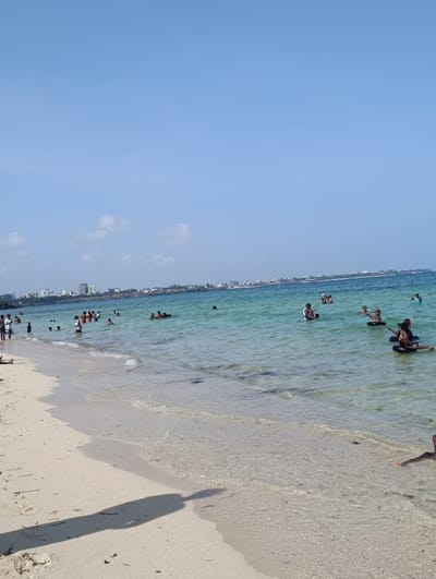 The image depicts a sunlit coastal scene in Makadara/Mji Wa Kale ward, Kenya, featuring a sandy beach and clear blue-green ocean water under a predominantly light blue sky with minimal cloud cover. Approximately 20-30 individuals, including adults and children, are present. Most people are observed in the water, wading, standing, or swimming; some are chest-deep, while others are closer to the shore. Two individuals on the right side are using black inflatable inner tubes. A few figures are visible further down the beach to the left, near the water's edge. The foreground features light-colored sand with scattered dark debris and a prominent elongated shadow extending from the lower-left towards the water. In the distant background, a coastline dotted with numerous white and light-colored urban buildings is visible along the horizon. The scene indicates recreational activity on a clear day.