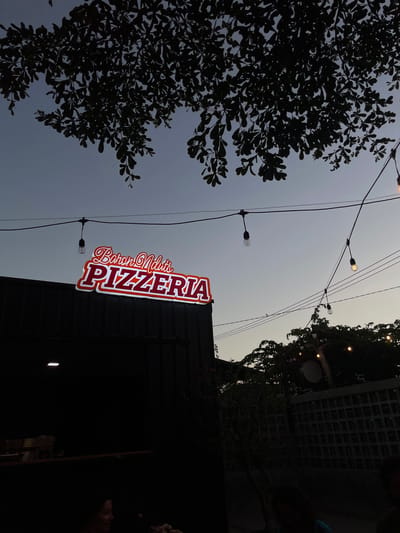 The image depicts an outdoor setting at dusk, with a neon sign for "Baron Nduts PIZZERIA" illuminated on the dark facade of a building. String lights with exposed bulbs hang across the scene, casting a dim glow. Silhouetted trees and foliage frame the sky, which transitions from a pale blue to a darker twilight hue. Part of a dark-colored structure, likely a storefront or restaurant, is visible on the left, with some items on a shelf. In the lower foreground, the silhouette of a person is visible on the left, and a dark area on the right suggests more of the outdoor seating or dining area. The overall ambiance is casual and inviting, suitable for an evening meal. This establishment is located in the Tamansari area of Indonesia.