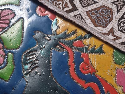 The image shows a close-up of a colorful, textured surface, possibly a metal plate or painted wood, with a fabric overlaying part of it. The surface beneath the fabric is adorned with stylized floral and mythical creature motifs rendered in bright, unevenly applied paint. Dominant colors include deep blue, black, red, yellow, green, and pink. The paint is chipped and weathered, revealing a metallic sheen and some rust in places. The texture is rough, with visible brushstrokes and small bumps. A piece of woven fabric with a geometric pattern in brown and cream is draped over the upper right portion of the painted surface. This appears to be a decorative element on an object, possibly a panel or a piece of furniture, located outdoors, possibly in a public space in Shopokov, Kyrgyzstan. No people are visible in the image.