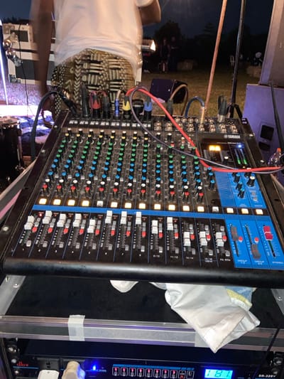 The image depicts an outdoor sound engineering setup at night in Murera ward, Kenya. A black Yamaha MG16XU mixing console occupies the foreground, positioned on a rack or stand. The console features numerous green, blue, and black rotary knobs, and multiple white faders. Several faders display illuminated red "PEAK" indicators, suggesting active audio signals. Various colored audio cables, including red and blue, are connected to the console's input/output ports. Directly beneath the console, a white cloth is visible, partially obscuring a rack-mounted dbx SR-328V audio processor, which displays "181" on its blue digital screen.

In the midground, partially visible behind the mixing console, stands an individual wearing a white t-shirt and patterned shorts featuring a black and beige geometric design. The person's stance suggests they are operating or attending to the audio equipment. The background reveals a dimly lit outdoor area with sparse vegetation or grass. Blurry figures are discernible in the distance, indicating other attendees or personnel at an event. Stage lighting elements, including a bright light source on the far left and a dark drum component, are present. A vertical brown pole or stand is visible on the right side of the frame. The overall scene suggests a live performance or public event setting.
