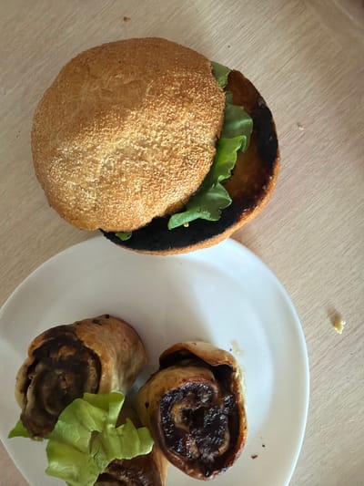 The image displays a close-up, overhead view of a meal on a light-colored wooden surface. The main subjects are a hamburger and several pastry rolls on a white plate.
The hamburger is partially visible, with its top bun on display. It appears to be a sesame seed bun, and some green lettuce is peeking out from underneath. The bottom bun and the patty are obscured. The top bun has a dark, slightly burnt edge.
To the right of the hamburger, on a white plate, are three pastry rolls. These are sliced to reveal a dark, jam-like filling, possibly fruit or chocolate. One of the rolls has a piece of lettuce placed on top of it. The pastries appear to be golden brown and flaky.
The wooden surface has a visible grain. There are a few small, light-colored crumbs scattered on the surface, one particularly noticeable near the top left and another near the top middle.
There is no visible text in the image. The lighting suggests it might be daytime, with natural light illuminating the scene from above or slightly to the side. The overall impression is that of a casual meal. The scene is set in Lopatino, Russia.