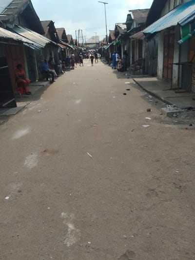 A street scene depicts an unpaved road lined with weathered, tin-roofed buildings. People gather along the edges, some sitting and others walking in the distance. Litter and debris are scattered on the road surface. The buildings show signs of age and wear, with faded paint and closed shutters. Overhead, a streetlight is visible in the distance. The overall impression is of a humble, lived-in environment.