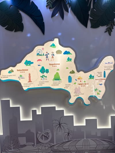 An indoor, illuminated wall display presents a stylized, three-dimensional map illustrating the districts of Sanya City, Hainan Province, China. The map, rendered in light tones with a prominent light blue lower border simulating a coastline, is centrally positioned and divided by dashed lines into distinct administrative areas, including Tianya District, Yazhou District, Jiyang District, and Haitang District.

Each district on the map features colorful, cartoon-style icons representing specific landmarks and attractions, accompanied by English and Chinese text labels with small locator pins. Tianya District, located towards the upper-center of the map, is depicted with a Ferris wheel, a parachutist descending, two stylized figures in local attire, and a traditional hut structure. Yazhou District, to the left of Tianya District, showcases icons for Sanya Dongtian Park, Nanshan Cultural Tourism Zone (a tall pagoda-like structure), Coconut Dream Corridor (palm trees), and Tianya Haijiao Scenic Spot (green rock formations). Jiyang District, situated below Tianya District, includes icons for Phoenix Island (a blue skyscraper-like building), a camping tent, and Luhuitou Park (a statue of a deer and figures). Haitang District, to the right, features icons for Sanya International Duty-Free Shopping Complex and Atlantis Sanya Resort (both represented by tall skyscrapers), and figures engaged in water activities.

Above the map, several layered, dark green, cutout shapes resembling tropical leaves are affixed to the wall, some illuminated by spotlights. Below the map, a multi-layered grey silhouette artwork depicts a modern cityscape, including numerous buildings, palm trees, and a large, stylized infinity symbol. This cityscape element is backlit along its upper edge, creating a glowing separation from the map above. The entire display is mounted on a uniform, light-colored wall, utilizing internal backlighting for the map and cityscape, along with external illumination for the foliage elements.