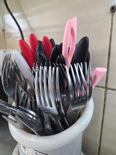 A white cylindrical utensil holder, possibly plastic or ceramic, is positioned on a dark, reflective surface, likely a countertop. The holder contains an assortment of clean cutlery. Numerous stainless steel forks are vertically arranged with their tines pointed upwards, largely filling the container. Interspersed among the forks are multiple knives featuring opaque plastic handles of various colors: several black, at least four red, one white, and two pink-handled knives. One of the pink-handled knives exhibits a distinct open-loop design integrated into its handle. Faint, partial text and a barcode are visible on the lower section of the holder. The background consists of a light-toned ceramic tiled wall with clearly visible grout lines. A black electrical cable descends vertically in the upper left quadrant of the background. A silver Phillips head screw is embedded within a horizontal grout line in the upper right quadrant. No specific identifying features are present in the image to determine a precise location within the city of Gevgelija, North Macedonia.