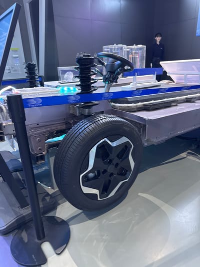 The image displays an indoor exhibition setting in Guangming District, China, featuring a demonstration cutaway of an electric vehicle chassis. The primary subject is a grey automotive frame, presented as a platform, with a single black tire and a black and silver five-spoke alloy wheel visible on the right side. Above the wheel, a black coil spring and strut assembly are mounted, adjacent to a partially visible black steering wheel and column. A second identical coil spring and strut assembly is positioned towards the left of the chassis. A blue banner, adorned with Chinese text and a logo, spans the mid-section of the display, running above a segment of blue LED lighting that illuminates transparent components within the chassis, suggesting internal electrical or battery elements. In the upper right background, one individual, a young female with dark hair, stands upright, dressed in a dark long-sleeved shirt and dark trousers, observing the exhibit without direct engagement. Transparent display units containing other components are visible behind the main chassis display. The environment consists of a light grey, reflective floor and dark grey walls, illuminated by artificial lighting. A black stanchion with a circular base is present in the lower-left foreground.