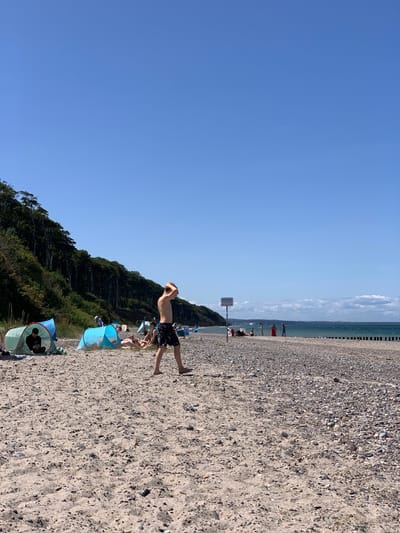 The image depicts a sunny day on the beach in Nienhagen, Germany. In the foreground, a young person in black and white patterned swim shorts walks across the sandy beach, with their hands raised to their head. Further back, several people are seen relaxing near colorful beach tents, including a green tent with a person sitting inside and a blue tent with people lying down. The beach transitions from sand to pebbles as it meets the sea. A line of wooden posts serves as a breakwater in the water. On the left side of the image, a steep, tree-covered cliff forms a backdrop to the beach. A sign is visible on the beach with German text, reading "BEGINN FISCHEREI-BEREICH". The sky is clear blue, with a few distant clouds.