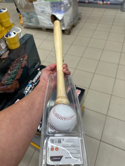 A person's arm and hand are holding a baseball bat and ball set in a retail environment. The baseball bat is made of light-colored wood and has a smooth finish. The baseball is white with red stitching. Both are packaged in clear plastic. In the background, there are shelves stocked with various products, including what appear to be rolls of tape and cardboard boxes, suggesting a store setting. The floor is tiled in a light color. The setting is consistent with a retail store in Orsk, Russia, given the product labeling and the store's general appearance.