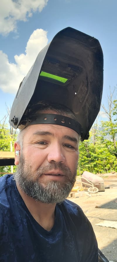 In an outdoor setting, a man with graying hair and beard is seen wearing a welding mask. The mask is dark and covers the top of his head, with a bright green lens visible. He has a grey wireless earbud in his left ear. He is wearing a dark blue t-shirt with a subtle patterned design. The background shows lush green trees and a hazy mountain range, suggesting a rural or semi-rural location. Sunlight illuminates the scene, casting shadows. There are some construction materials or equipment visible in the background, including what appears to be a wheelbarrow. The sky is blue with scattered white clouds. The location is identified as Milisi, Georgia.