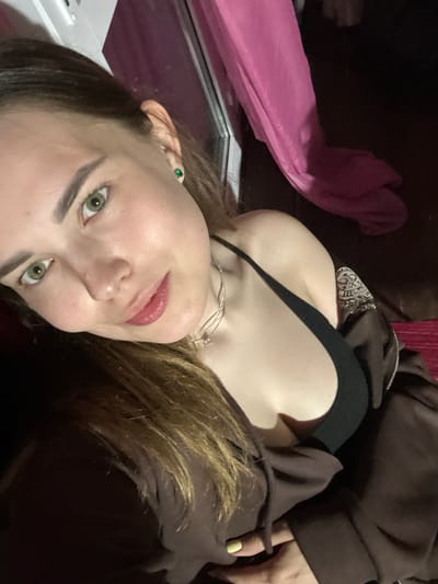 A young woman with green eyes and brown hair is taking a selfie indoors. She is wearing a black camisole and a brown zip-up hoodie. A pink fabric is visible in the background, along with what appears to be a window frame. The lighting is dim, suggesting it might be evening or nighttime. There is no information in the image to suggest a specific location within Zhebrei, Russia.