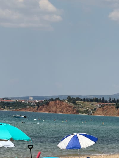 The image shows a view of the sea and a beach on a clear day, with a town visible in the distance. In the foreground, two beach umbrellas are set up on the sand, one turquoise and white, and another blue and white. The turquoise umbrella is in the lower left, and the blue and white umbrella is in the lower right. The sea is calm and a moderate blue color. A small white boat with a black outboard motor is visible in the water, to the left of center. A person is swimming in the water near the boat. Several buoys are also scattered in the water.

In the background, a reddish-brown cliff face dominates the coastline. On top of the cliff, there are buildings, trees, and a road. Further in the distance, a city skyline is visible with several buildings, including a prominent, tall, light-colored rectangular structure. This scene is located in Πόρτες, Greece. The sky is light blue with a few scattered white clouds. The overall impression is of a sunny day at the beach.