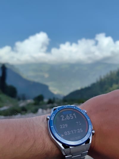 The image shows a close-up of a person's wrist wearing a silver smartwatch with a blue bezel. The smartwatch display shows "2,651" in large numbers, likely representing steps taken, along with other data including "229", "76", "03:17:25", and "16:07". The watch is on a person's forearm, which is covered in fine hair. The background is a blurred landscape of mountains and sky with clouds, suggesting an outdoor location. The provided crops indicate the background features are indeed a natural, mountainous environment. The image is taken from a low angle, focusing on the smartwatch. The location is identified as Palchan, India.