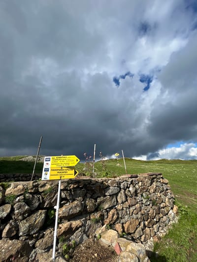 This is a vertical, eye-level, medium shot of a mountain trail signpost in the city of Rozino, Bulgaria. The signpost is located on a rocky embankment. The signpost has two yellow directional arrows with text in Bulgarian. The top arrow points to the right and indicates "Vr. Yumruka (1819 m.a.s.l.) 1.55km - 0.30h +170/-10m PARAKLIS SV. TROITSA - 0.10h Chapel St. Trinity". The bottom arrow also points to the right and reads "PANORAMEN KRGOV UKAZATEL PANORAMIC CIRCULAR INFORMANT 90 m". In the background, there is a grassy, rolling hillside under a dramatic sky filled with dark, stormy clouds with patches of blue sky peeking through.