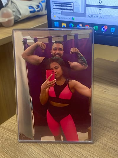 A photograph in a clear plastic frame shows a man and woman in workout clothes posing for a selfie in front of a purple background. The man is flexing his arm; he has a tattoo on his upper arm. The woman is holding a red phone and taking the selfie. The photograph and a laptop are resting on a wooden surface. The laptop displays icons and software windows. A white package with blue lettering is visible. The location is not discernible from the image. No evidence indicates the city of Luzhitsy, Russia.