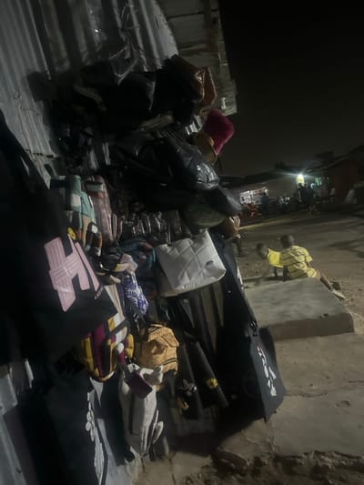 A cluttered scene displays numerous bags and luggage strewn on a corrugated surface outdoors. The bags vary in color, style, and material, including black, white, yellow, and patterned designs. Some appear to be handbags, while others are larger travel bags. To the right, a figure in a yellow shirt sits on a ledge. The background suggests an urban environment at night, with dimly lit buildings visible in the distance.