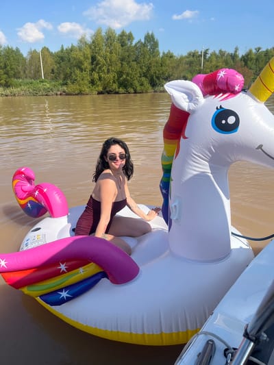 A woman is lying on an inflatable unicorn in the water. The woman has dark hair and is wearing a maroon swimsuit and sunglasses. She is smiling at the camera. The inflatable unicorn is white with a pink and yellow mane and tail. The water is brown and the sky is blue with white clouds. Trees line the shore in the background. The location is San Fernando, Argentina.