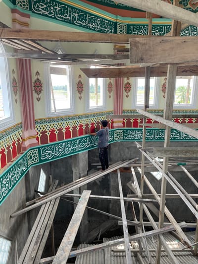 An interior view depicts an ongoing construction or renovation project within a building in Mns Asan, Indonesia. A male individual, wearing a grey short-sleeved shirt and paint-splattered dark pants, stands barefoot on a horizontal wooden plank. He is actively engaged in painting a red arch-shaped element on an elaborate, multi-colored decorative frieze on a wall.
The room's upper walls are adorned with light yellow panels featuring symmetrical red, pink, and green floral-geometric patterns, interspersed with arched windows framed in white. A continuous band of white Arabic calligraphy on a dark green background extends horizontally below these panels. Beneath the frieze being painted, another band of white Arabic calligraphy is present on a lighter green base, above unfinished grey concrete walls. The lower section of the image is dominated by a complex network of rough-hewn wooden planks and beams forming scaffolding, some resting on corrugated metal sheets on the floor below. Through the multiple windows, a bright exterior is visible, showing expansive green fields, distant trees, and a palm tree under a bright sky.