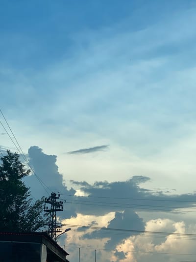 This is an outdoor, eye-level, vertical medium shot of a cloudy sky with electrical wires and a utility pole. The sky is a light blue with scattered white and gray clouds, some of which appear dark and textured. In the lower left corner, the dark green foliage of a tree is visible. A dark, silhouetted structure, possibly a building, is in the bottom left, with a portion of its roof showing red trim lights. A black utility pole with insulators and wires stretches up into the sky from the lower center, supporting multiple black electrical wires that extend horizontally across the frame. A street lamp is attached to the utility pole, pointing downwards. In the distance, on the right side of the frame, the sky transitions to a lighter, more yellowish hue, suggesting the sun may be setting or obscured by clouds. The image appears to be taken in Zrenjanin, Serbia, based on the context provided. There are no people visible in the image.