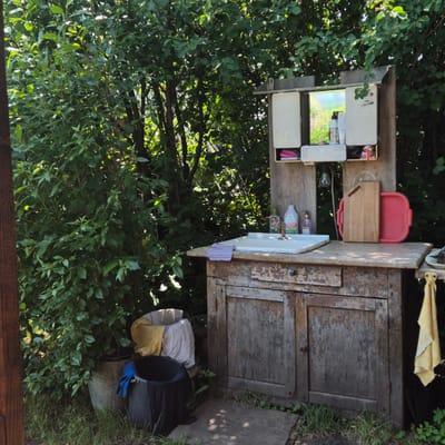 This is a medium shot, eye-level, outdoor photograph of a rustic outdoor sink setup located amidst lush green foliage in Новостройка, Russia. The sink is part of an old, weathered wooden cabinet with peeling paint, suggesting it's in a rural or garden setting. Above the sink, there's a small mirrored cabinet with its doors open, revealing various items inside, including bottles of soap or lotion, and a pink slipper. A light bulb hangs from the cabinet. On the countertop, next to the sink, are two bottles of cleaning liquid, one white and one purple, and a faucet. A wooden cutting board and a red plastic tray are positioned to the right of the sink. In front of the cabinet, two buckets are visible; one is a larger grey metal bucket containing laundry, and a smaller black plastic bucket with blue gloves and dark fabric. The ground around the sink is covered in grass and dirt, with some fallen leaves. The background is dominated by dense green bushes and trees, indicating a natural, possibly overgrown environment. Sunlight filters through the leaves, creating dappled shadows on the ground and the sink area. The scene is static, with no people present, and appears to be a functional, albeit unkempt, outdoor washing station.
