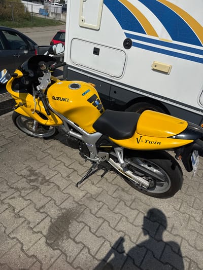 A yellow Suzuki SV650S motorcycle is parked on a paved area in front of a white and blue recreational vehicle. The motorcycle is angled towards the right, showing its front wheel, handlebars, fuel tank, and seat. The fuel tank has "SUZUKI" branding and a "DENESE" sticker. The rear of the motorcycle displays "V-Twin" branding and a license plate that appears to be from Germany, with the letters "S" and "TU" visible. The RV has a blue and yellow striped decal on its side. The ground is made of interlocking gray paving stones, with some dark stains that could indicate oil or water. In the background, a dark colored car and a red car are visible. The scene suggests an outdoor setting, possibly a parking lot or campsite, with a residential or commercial building in the far background. The overall lighting indicates it is daytime with bright sunlight casting a shadow of a person on the ground. The image was taken in Wettstetten, Germany.