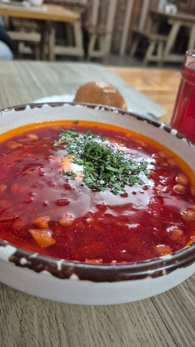 The image shows a close-up, high-angle view of a bowl of borscht, a traditional Eastern European soup. The soup is a vibrant red color, indicating the presence of beets, and contains visible chunks of vegetables. A dollop of white cream or sour cream is in the center, topped with a generous garnish of fresh green herbs, likely dill or parsley. The bowl itself has a rustic appearance with a white ceramic base and a dark, possibly brown, decorative rim.
The soup is placed on a light brown wooden table, suggesting a casual dining setting. To the left of the bowl, partially visible, is a round, golden-brown bread roll, often served with borscht. In the background, out of focus, are elements that suggest an indoor environment, possibly a restaurant or home kitchen. There are glimpses of what appear to be wooden furniture or shelves, and a hint of a red liquid in a bottle or glass, possibly a beverage to accompany the meal.
The lighting is soft, suggesting it could be daytime, but the exact time of day is not discernible. There are no discernible weather cues. The overall impression is one of a hearty, home-cooked meal being served. The focus on the soup and its garnishes suggests an appreciation for the food. No text is visible in the image. The location context provided is Bol'shaya Glubokaya, Russia, which aligns with the type of cuisine depicted.