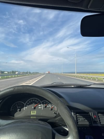 The image presents a view from inside a car, rotated 90 degrees clockwise from a standard forward perspective. The right side of the frame is occupied by the car's dark interior, including a portion of the steering wheel and the instrument cluster. The left side features a bright blue sky with scattered white, wispy clouds, seen through the windshield.

The main subject is a long, straight road extending vertically into the distance, visible through the windshield. The road appears to be a highway or a bridge, with a grey guardrail running along its left side. Multiple white light poles line the road, receding towards the horizon. A small, distant vehicle, possibly red, can be seen far ahead on the road. Beyond the guardrail, a flat landscape, possibly fields or water, stretches towards the distant horizon. The overall setting suggests travel on an open road in Усть-Сыны, Russia, likely near the Krasnoyarsk Reservoir.

The car is in motion, as indicated by the speedometer. Visible details on the dashboard include a black steering wheel with a textured grip, and an instrument cluster featuring a speedometer, tachometer, and digital display. The speedometer needle points to approximately 80 km/h. The digital display shows "190901" (likely odometer or trip mileage) and "18:26", indicating the time is 6:26 PM. The weather is clear and sunny. A red triangular hazard light button and air vents are also visible on the dashboard.