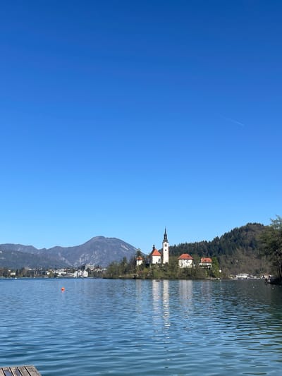 The image depicts a scene at Lake Bled, Slovenia. A church with a tall steeple is situated on an island in the lake. A mountain is visible in the background. The lake's water surface is rippled. The sky is clear and blue. A visible contrail from a passing aircraft crosses the sky. A forest exists on the edge of the lake.