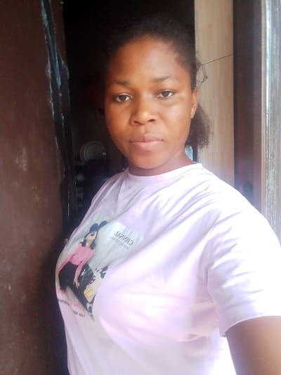 This is a medium shot photograph of a young woman with brown skin and dark hair, looking directly at the camera. She is wearing a light pink t-shirt with a graphic print on the left side of her chest. The graphic appears to be a fashion illustration of a woman in a pink top and black skirt, alongside cartoon characters. Some text is visible on the t-shirt, appearing to be reversed or mirrored, possibly "CHINVA" or a similar word.
The woman is standing in what appears to be an indoor setting with a slightly cluttered background. To her left is a dark, textured surface, possibly a wooden door or wall. To her right, a lighter colored wall with vertical lines is visible, suggestive of wooden paneling or blinds, with a bright light source to the far right, indicating an opening like a window or doorway. In the background, on the left side, some stacked plates and other items are partially visible, suggesting a domestic environment.
The lighting suggests it is daytime, with natural light illuminating the scene from the right. The woman's expression is neutral and direct. The overall impression is that of a casual, candid photograph taken indoors, possibly in Yenegoa, Nigeria, given the provided location context.