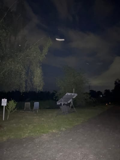 This is a night photograph taken in the city of Hanau, Germany. The scene depicts an outdoor area with a grassy ground and a dirt path in the foreground. In the middle ground, there are two chairs, a small bush, and a swing set. A signpost is visible to the left of the chairs. The background features trees and a dark sky with clouds. A bright, elongated streak is visible in the sky, possibly indicating a moving object or light artifact. The overall lighting suggests it is nighttime, with ambient light from the sky and potentially distant artificial sources.