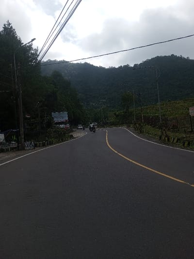 A road curves uphill with a yellow line dividing lanes. A motorcycle travels along the road. Lush green hills form the backdrop, partially obscured by a cloudy sky. Power lines stretch across the upper part of the image. Vegetation and sporadic streetlights line the roadside. A billboard and signage are visible on the left side of the road.