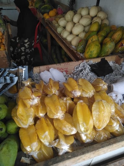 In a fruit market stall located in Yae Tha Yauk, Myanmar, a diverse array of fresh produce is displayed. The foreground features a large pile of approximately 30-40 bright yellow star fruits (carambola), each individually wrapped in clear plastic film, resting in a wooden crate lined with crumpled newspaper. To the immediate left of the star fruits, several green, spherical fruits, consistent with guavas or unripe mangoes, are partially visible.
Above and behind this display, an elevated wooden stand presents additional fruits. This stand holds a significant quantity of light-colored, oval melons, resembling honeydew or pale cantaloupes, concentrated in the center-right. Adjacent to these melons on the right, a stack of green and yellow papayas is arranged, some also appearing to be plastic-wrapped. To the left of the melons, smaller orange fruits (likely oranges) and larger green fruits (watermelons) are visible. A single pineapple stands upright at the upper-left edge of this elevated display.
In the upper-left background, a person is partially visible from the back, wearing a dark garment and a black head covering. They are bent over, attending to items on a lower display surface. A red plastic chair is positioned behind and to the right of the individual. The overall environment suggests a bustling market setting with various fruits openly displayed on wooden structures.