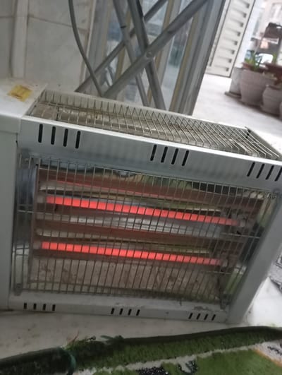 An electric space heater, white in color with a metallic front grate, is depicted in an active state, indicated by four glowing red heating elements. Dust and debris are visible on the internal reflective surface behind the elements. The heater rests on a light-colored, flat surface. A green patterned rug is partially visible in the lower foreground. In the background, multiple leaning glass panels or window frames are visible. Beyond these, an outdoor area with a concrete or tiled surface displays several light-colored plant pots, some containing green plants, and a white, vertically slatted structure. The scene is located in the city of Nasiriyah, Iraq.