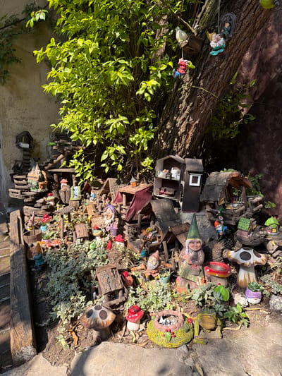 The image depicts a whimsical garden display in Domingo Arenas, Mexico. The scene features an assortment of garden gnomes and miniature structures, including a multi-level miniature home with visible interior details. There are also ceramic mushrooms and various small objects placed amidst greenery, including low-lying shrubs and climbing vines. A tree trunk is visible to the left. The composition is arranged in a somewhat haphazard manner, suggesting a playful and imaginative design. The environment appears sunny, with shadows cast across the scene.